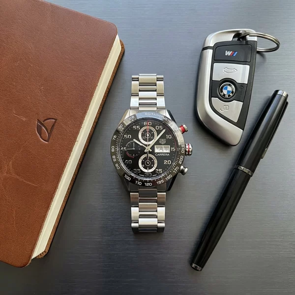 TAG Heuer Carrera Chronograph placed on dark brushed aluminium surface alongside a leather notebook a set of car keys and a premium pen shot from above