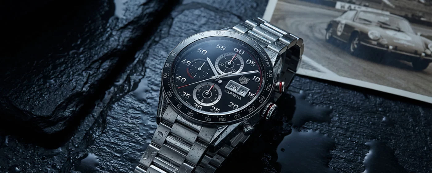 TAG Heuer Carrera Chronograph with black dial lying at a slight angle on dark wet slate with a blurred vintage racing photograph visible in the background