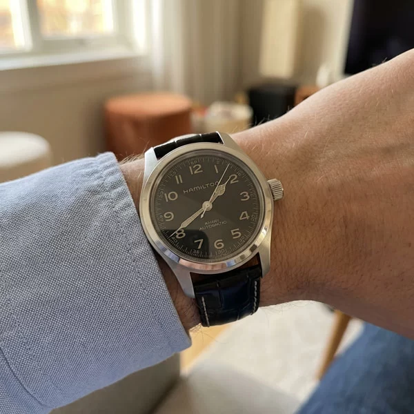 Hamilton Khaki Field Murph 38mm watch with black dial and black leather strap worn on a wrist with a light blue dress shirt sleeve, photographed from above in warm indoor natural light with a blurred living room interior in the background