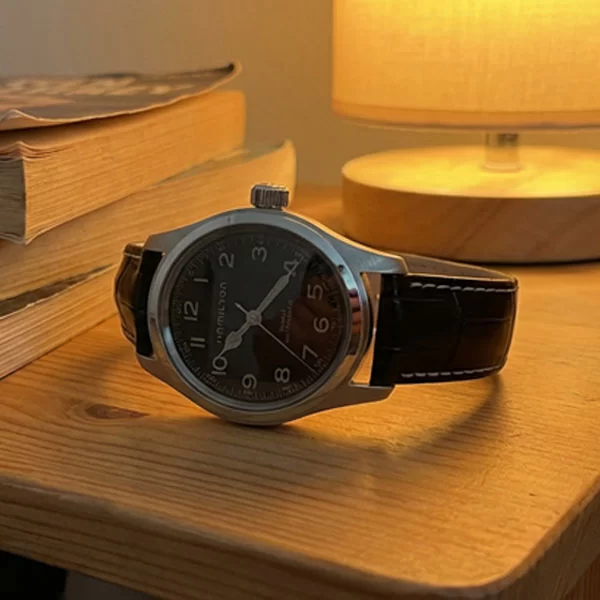 Hamilton Khaki Field Murph 38mm watch with black dial and brown leather strap placed on a wooden bedside table next to a stack of old paperback books, with a round table lamp glowing warmly in the background