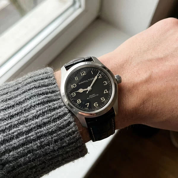 Hamilton Khaki Field Murph 38mm watch with black dial and black leather strap worn on a wrist with a grey knit sweater sleeve, photographed from above near a window in natural light