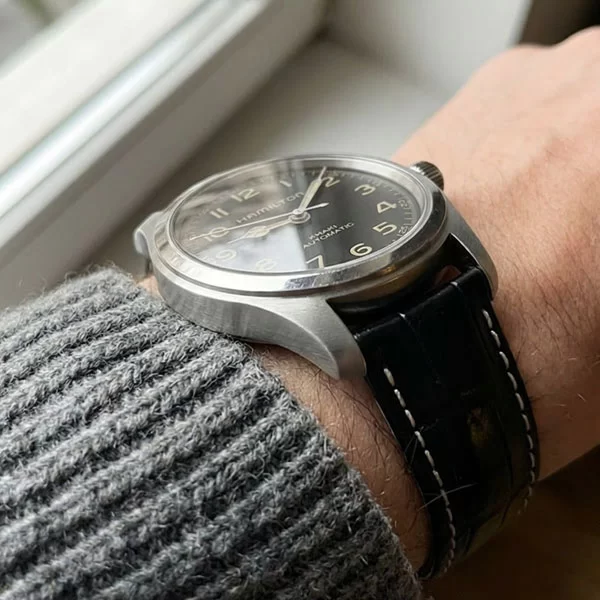 Hamilton Khaki Field Murph 38mm watch with black dial and black leather strap with white contrast stitching worn on a wrist with a grey knit sweater sleeve, photographed from the side at a low angle showing the case profile and lug area