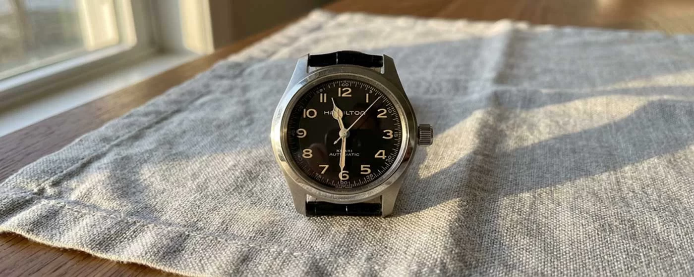 Hamilton Khaki Field Murph 38mm watch with black dial, beige Arabic numerals, and cathedral hands resting upright on a grey linen cloth on a wooden table, natural window light casting soft diagonal shadows across the surface