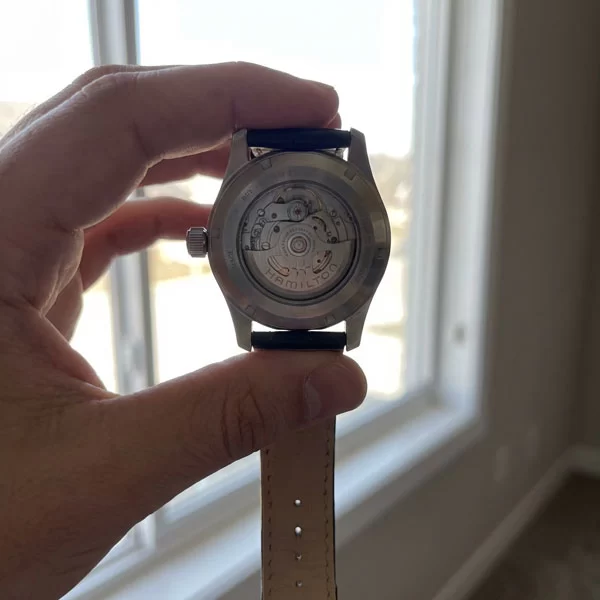 Hamilton Khaki Field Murph 38mm held in a hand facing caseback up toward a bright window, the exhibition sapphire glass revealing the H-10 automatic movement with rotor and bridges visible, black leather strap hanging down