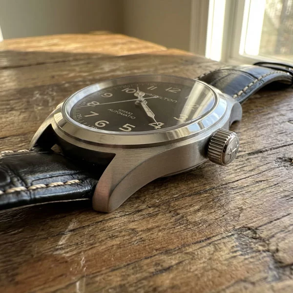 Hamilton Khaki Field Murph 38mm resting on a wooden surface at a low angle, showing the right side of the stainless steel case with the oversized push-pull crown, black leather strap with white contrast stitching visible, black dial partially visible from the front