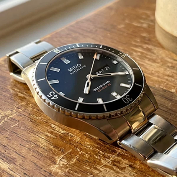 Mido Ocean Star 200 black dial stainless steel watch with bracelet resting on a worn wooden ledge near a window, ceramic mug blurred in the background, day-date display visible on the dial