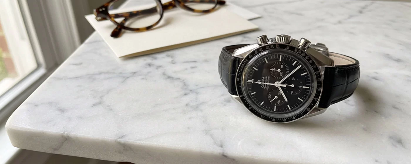 Omega Speedmaster Professional Moonwatch with black alligator leather strap resting on a marble surface next to tortoiseshell glasses