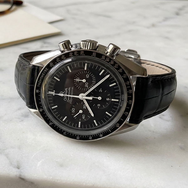 Omega Speedmaster Professional Moonwatch with black alligator leather strap resting on a marble surface next to tortoiseshell glasses