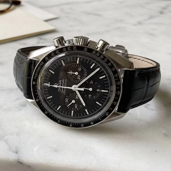 Omega Speedmaster Professional Moonwatch with black alligator leather strap resting on a marble surface next to tortoiseshell glasses
