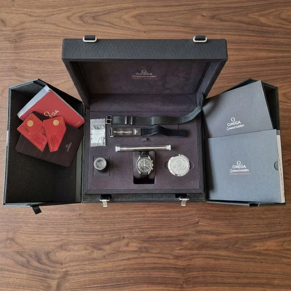 Omega Speedmaster Professional Moonwatch presentation box open on a walnut surface showing the watch, NATO strap, Velcro strap, caseback paperweight, and accessories