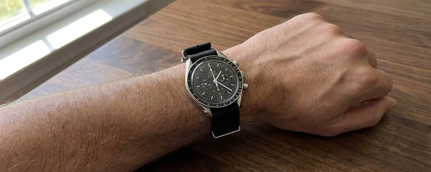 Omega Speedmaster Professional Moonwatch worn on a black NATO strap on the wrist resting on a wooden table