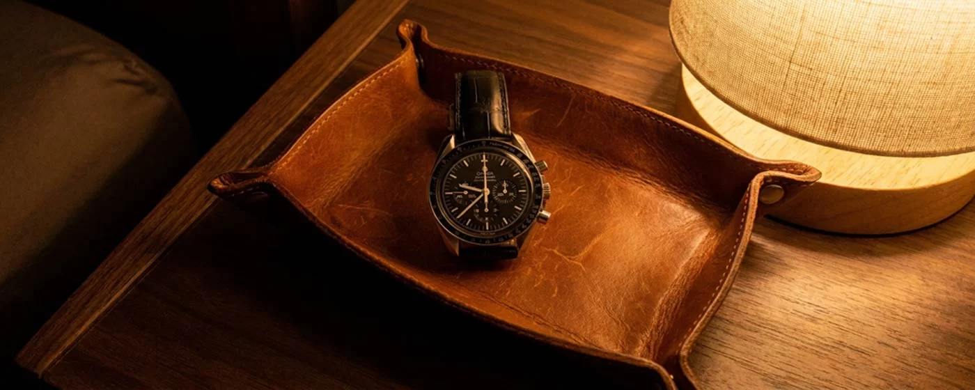 Omega Speedmaster Professional Moonwatch with black alligator leather strap resting in a cognac leather valet tray on a wooden surface beside a warm bedside lamp